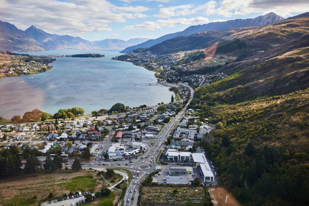 Aerial view of Queenstown, New Zealand, showing the lakefront town, surrounding homes, and mountain ranges under a blue sky.